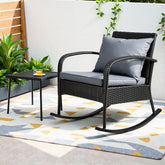 Rattan rocking chair with gray cushion on a patio with a small table and plants.