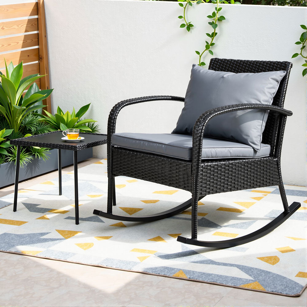 Rattan rocking chair with gray cushion on a patio with a small table and plants.
