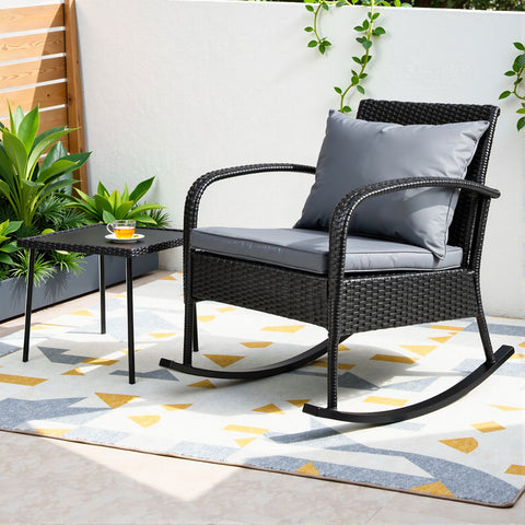 Rattan rocking chair with gray cushion on a patio with a small table and plants.